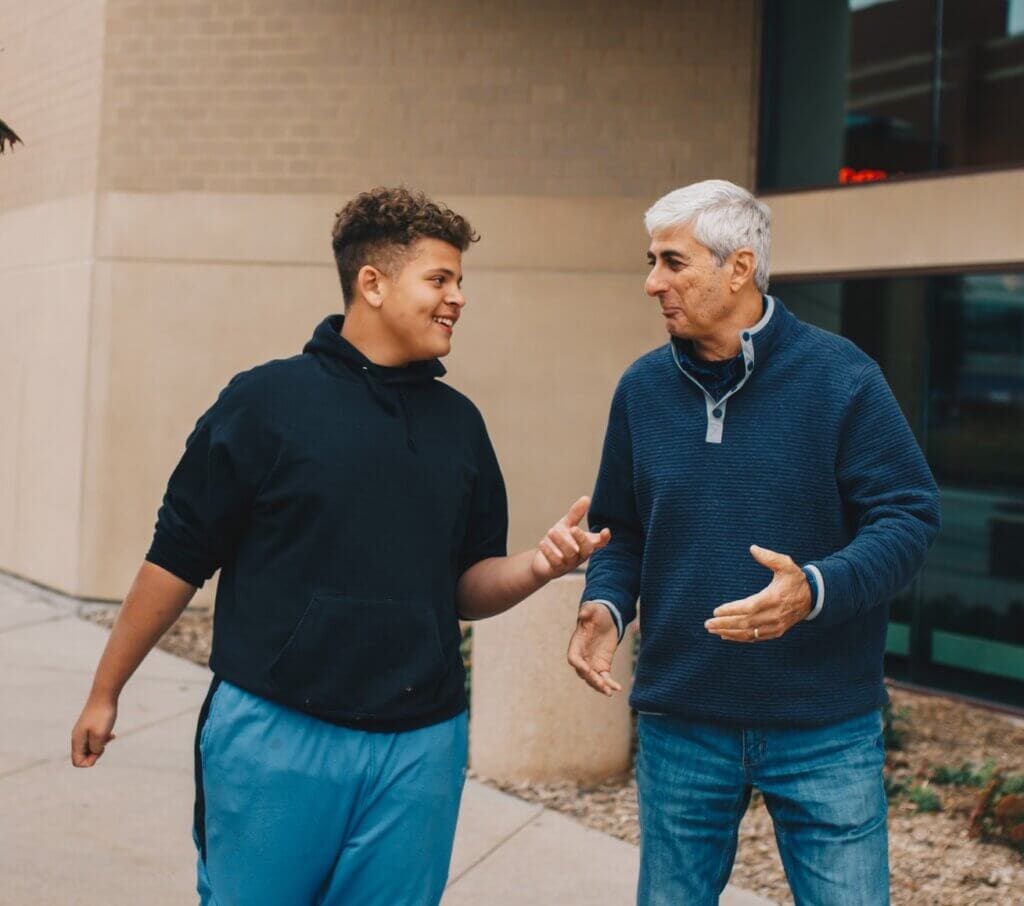 Brown haired teen male talks with white haired older male outside and they are smiling.